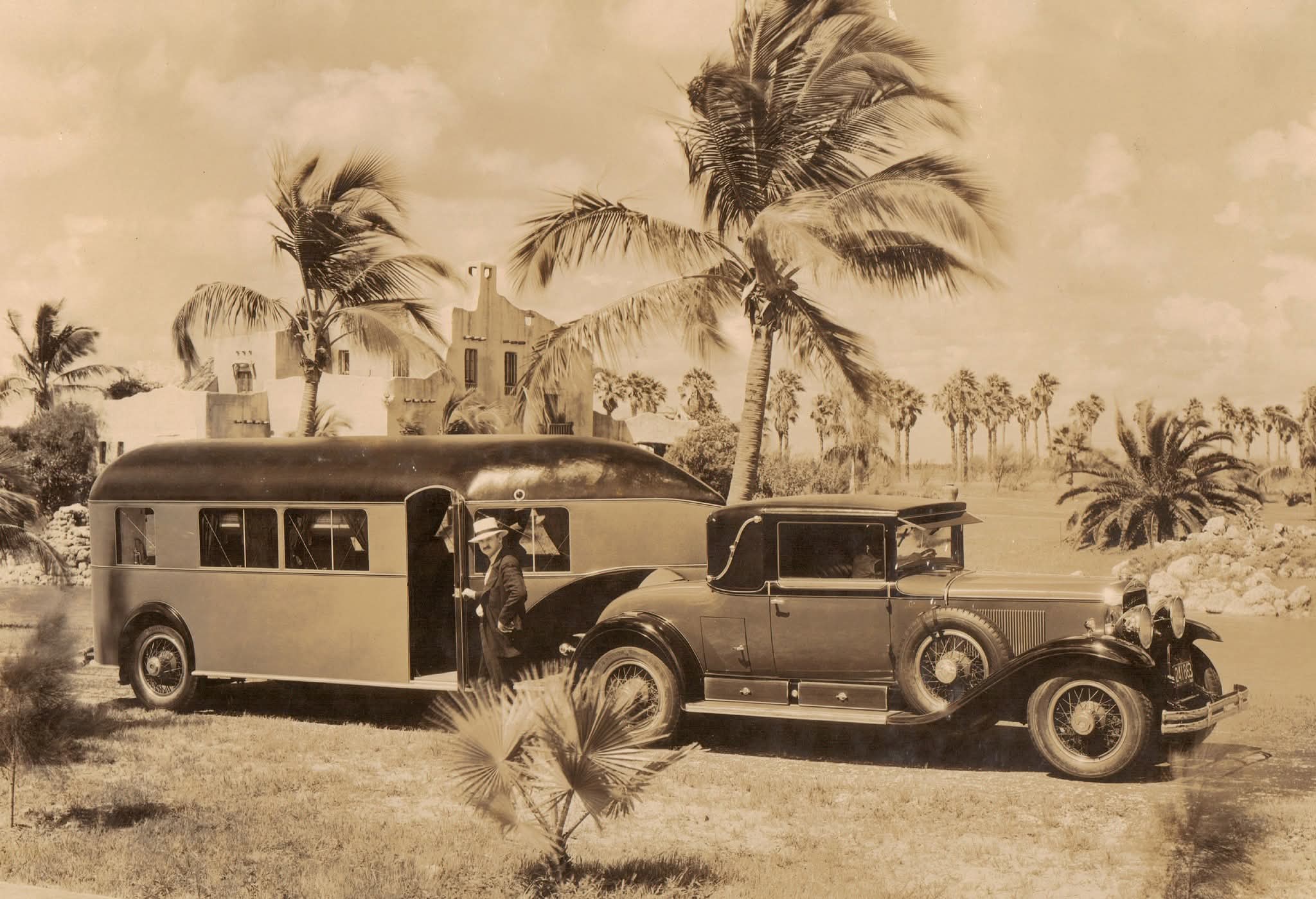 Un automóvil antiguo y una casa rodante estacionados frente a un edificio de estilo Pueblo Revival entre palmeras en los primeros años de Miami Springs